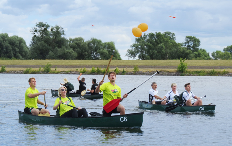 Foto: 3 Boote im Wettstreit, im vorderen Boot versucht ein Sportler stehend einen orangenen Luftballon über sich zu zerstechen. Foto: 3 Boote im Wettstreit, im vorderen Boot versucht ein Sportler stehend einen orangenen Luftballon über sich zu zerstechen.