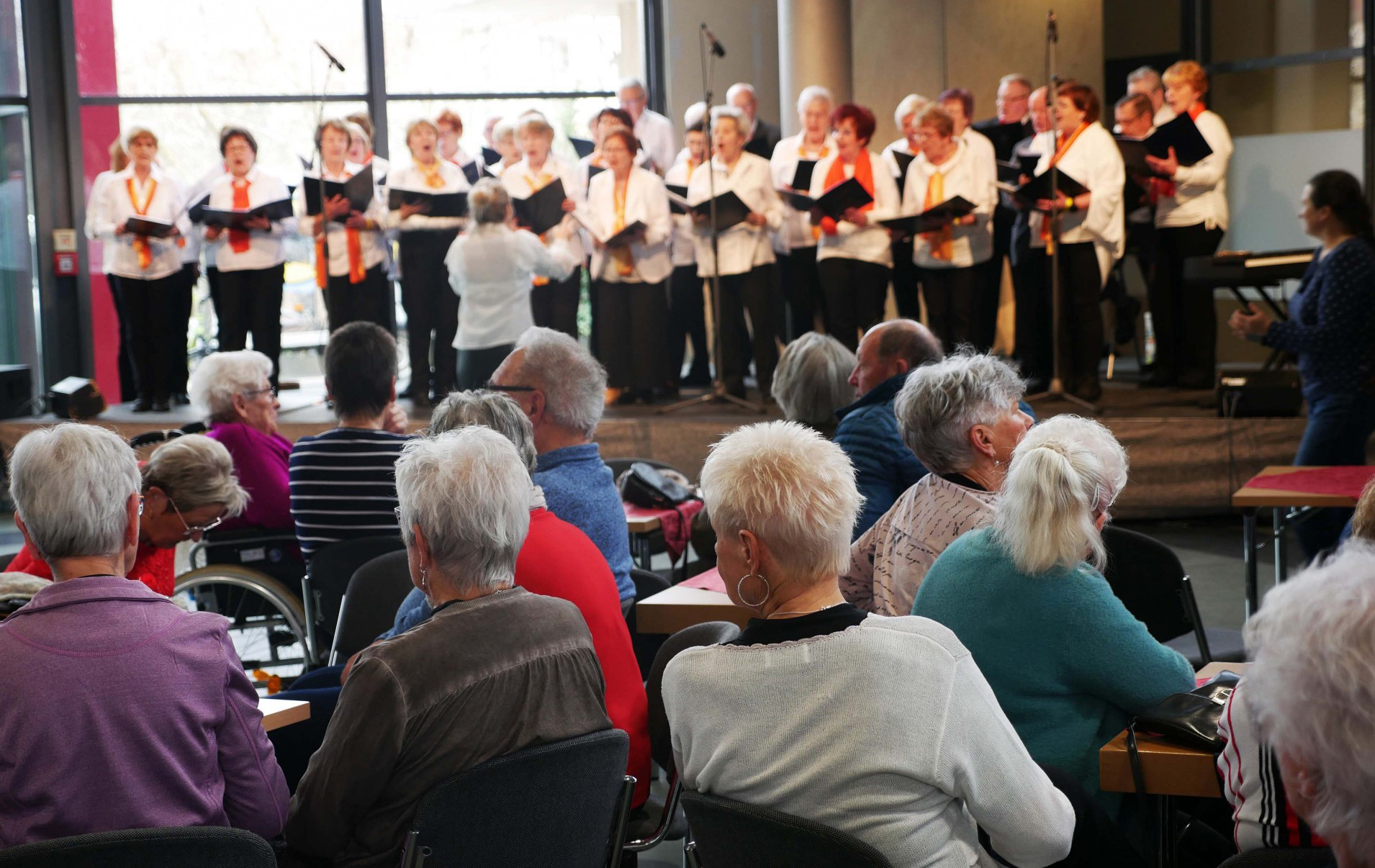 Foto: im Vordergrund grauhaarige Zuschauer von hinten, im Hintergrund unscharf ein Chor
